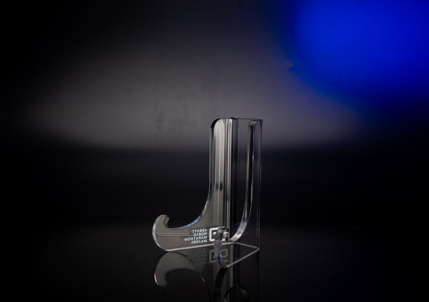 Clear acrylic display stand viewed from the right side, featuring curved supports and the Abbott World Marathon Majors logo, set against a dark background with a blue light gradient.