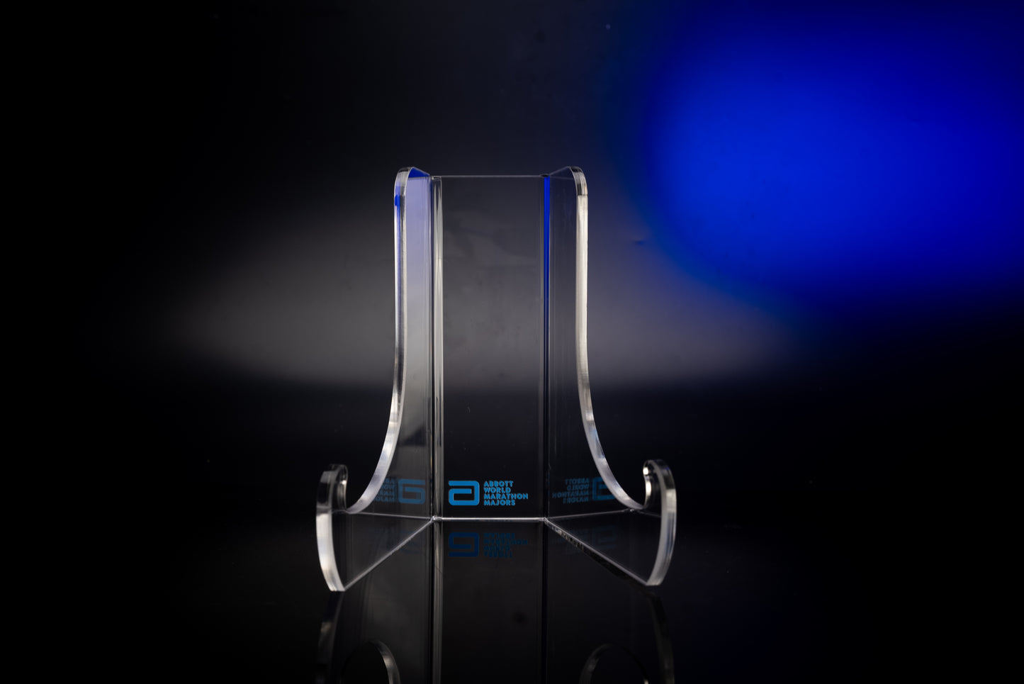 Front view of a clear acrylic display stand with curved base supports and the Abbott World Marathon Majors logo, photographed on a dark background with blue lighting.